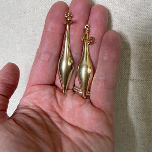 May include: A pair of gold-colored earrings. Each earring has a long, slender, teardrop-shaped body with a decorative top. The earrings are displayed on a neutral background.