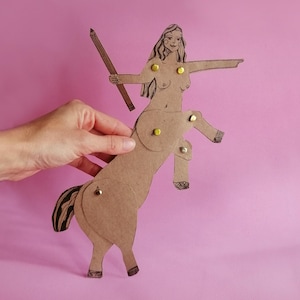 Può includere: Una figura di centauro di cartone fatta a mano con una matita, su sfondo rosa. Il centauro ha la parte superiore del corpo di una donna e la parte inferiore di un cavallo, collegati con borchie gialle. La figura è realizzata in cartone marrone con arte lineare nera.