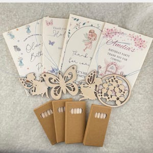 May include: Four rectangular cards with fairy and floral illustrations, each featuring names like Olivia and Amelia, paired with four light-coloured wooden cutouts in butterfly, fairy, and floral shapes, and four small brown cardboard boxes.