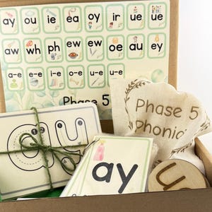 May include: A Phase 5 phonics educational kit in a cardboard box containing a reference chart with letter combinations, a stack of tracing cards tied with green twine, a ring of flashcards featuring the letters ay, and a small beige fabric bag.