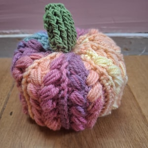 May include: A handcrafted, crocheted pumpkin in shades of pink, orange, and yellow, with a green stem. The pumpkin has a textured, braided design, and is sitting on a wooden surface. This decorative item is perfect for autumn.