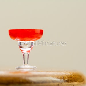 May include: A miniature red glass margarita glass on a wooden surface.