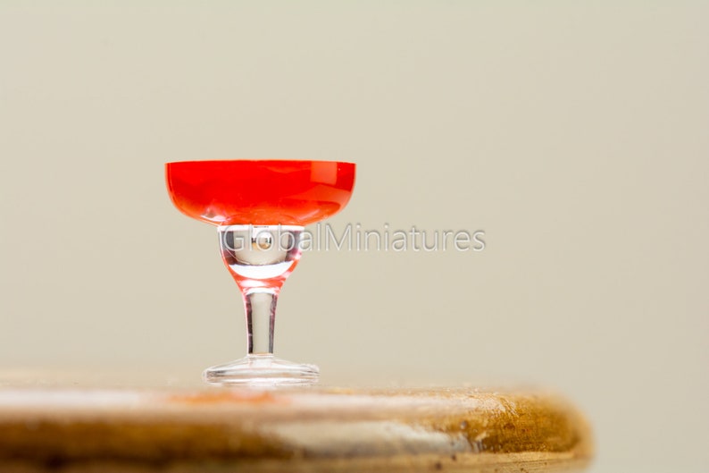 May include: A miniature red glass margarita glass on a wooden surface.