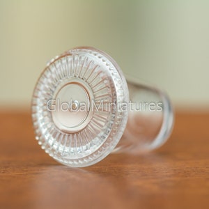 May include: A clear glass bottle stopper with a decorative, fluted rim. The stopper is lying on a wooden surface.