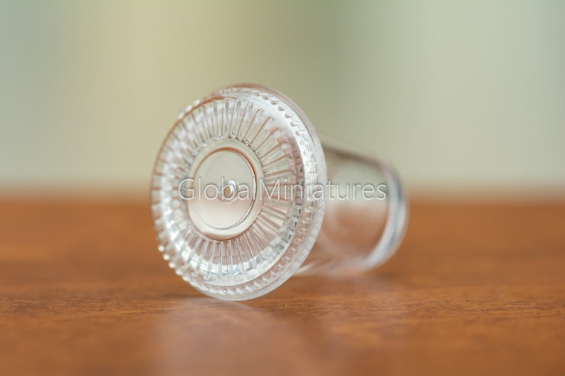 May include: A clear glass bottle stopper with a decorative, fluted rim. The stopper is lying on a wooden surface.
