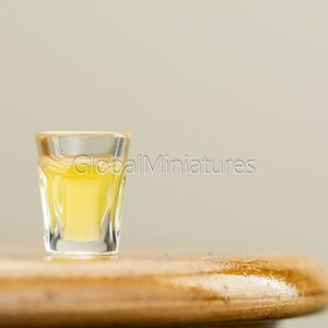 May include: A small shot glass filled with a clear yellow liquid sits on a wooden surface.