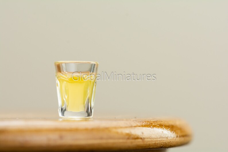 May include: A small shot glass filled with a clear yellow liquid sits on a wooden surface.