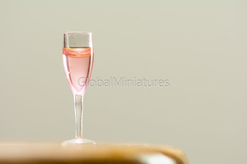 May include: A miniature glass filled with pink liquid, resembling a champagne flute. The glass is sitting on a wooden surface.