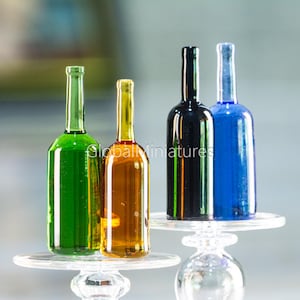Puede incluir: Miniaturas de botellas de vidrio en verde, ámbar, negro y azul, cada una llena de un líquido coloreado. Las botellas se exhiben sobre soportes decorativos transparentes, lo que sugiere una colección o exhibición de pequeños objetos decorativos.