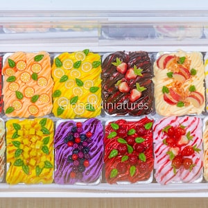 Puede incluir: Una vitrina llena de postres en miniatura coloridos. Los postres están dispuestos en bandejas rectangulares y cubiertos con varias frutas y adornos, incluyendo cerezas, naranjas, limones, fresas y kiwis. El texto "Global Miniatures" es visible.