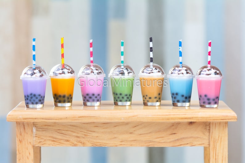 Puede incluir: Ocho vasos de pl&aacute;stico en miniatura con pajitas y tapas de colores, llenos de l&iacute;quidos de diferentes colores y perlas de boba. Los vasos est&aacute;n dispuestos sobre una mesa de madera.