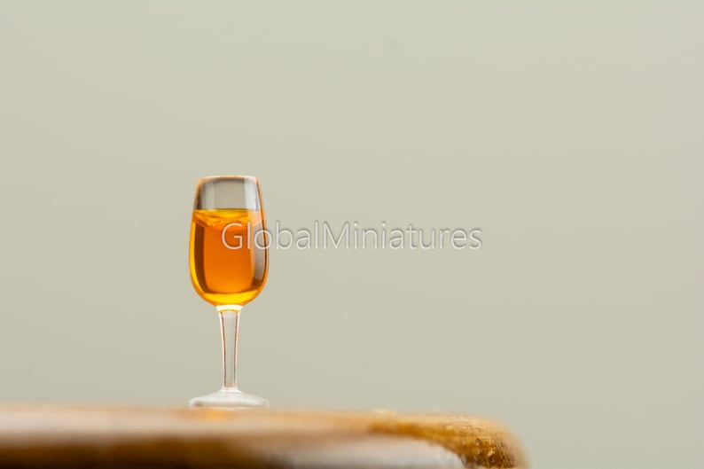 May include: A miniature glass of orange liquid sits on a wooden surface. The glass is clear and has a stem.