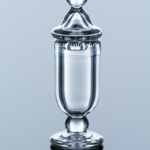 May include: A transparent glass apothecary jar with a rounded base, a cylindrical body, and a conical lid topped with a small sphere. The jar reflects light, and the text "tuasiwatn store" is printed on the body.