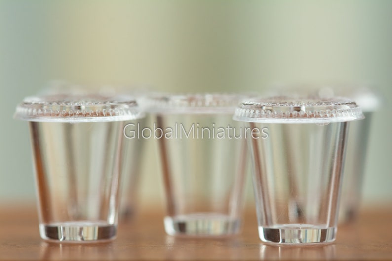 May include: Four clear plastic cups with lids, arranged in a row on a wooden surface. The cups are empty and appear to be made of a thin, transparent plastic.