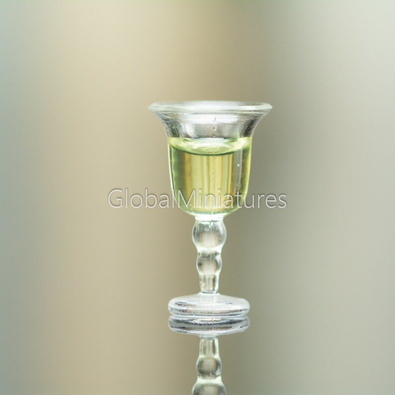 May include: A clear glass goblet with a round base and a stem with a series of small spheres. The goblet is filled with a light yellow liquid.