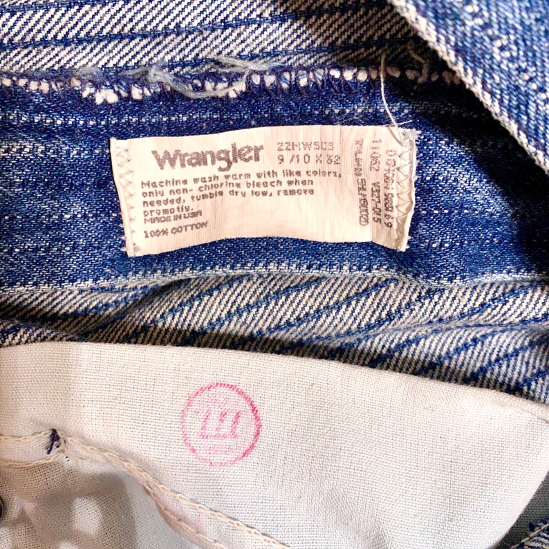 May include: A blue denim Wrangler jeans label with white text that reads "Wrangler 22HWS03 9/10 X 82 Machine wash warm with like colors, only non-chlorine bleach when needed, tumble dry low, remove promptly. Made in USA 100% COTTON".