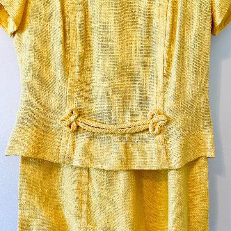 K&ouml;nnte beinhalten: Ein gelbes Leinenkleid mit kurzen &Auml;rmeln und einer taillierten Taille. Das Kleid hat ein einzigartiges Design mit einem Seildetail und geknoteten Akzenten.