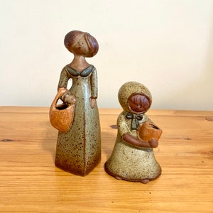 May include: Two ceramic figurines of women, both wearing dresses and carrying baskets. The taller figurine has brown hair and a green dress, while the shorter figurine has a brown hooded dress.