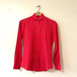 May include: A bright red long-sleeve blouse with a pleated front and a rounded collar. The blouse is buttoned down the front and hangs on a wooden hanger. The fabric appears to be a lightweight material.
