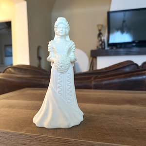 May include: A white ceramic figurine of a woman in a long dress, holding a bouquet of flowers. The figurine has detailed features, including a high collar and a decorative pattern on the dress. The figure is standing on a wooden surface.