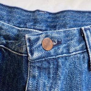 May include: Close-up of a blue denim waistband with a copper button. The button has a raised design.