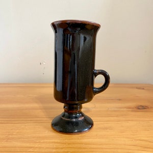 May include: A black ceramic mug with a brown rim and a handle. The mug has a pedestal base.