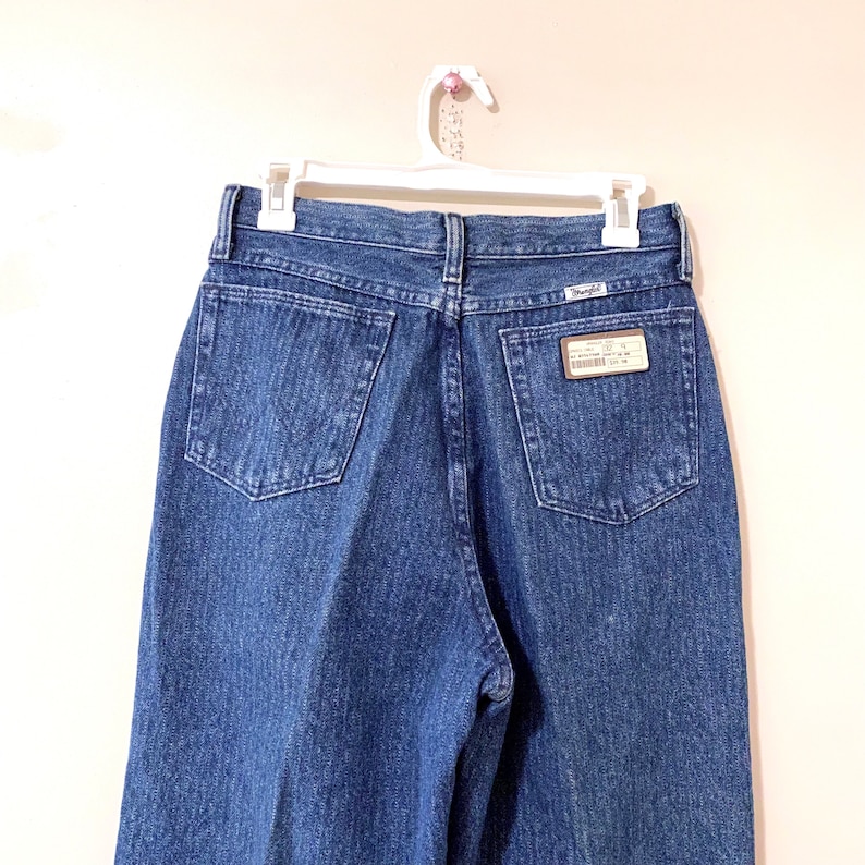 May include: A pair of blue denim jeans with a back pocket and a Wrangler label. The jeans are hanging on a white hanger.