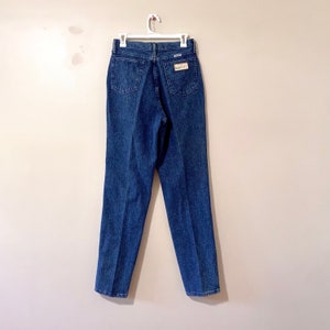 May include: A pair of dark blue denim jeans with a high waist and a straight leg. The jeans have a small brown leather patch on the back right pocket with the text "Wrangler" on it.