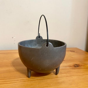 May include: A small, gray cast iron cauldron with a black wire handle and three legs. The cauldron is sitting on a wooden surface.