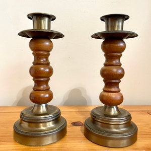 May include: A pair of brass and wood candle holders. The holders have a round base and a tall, slender column with a series of decorative wooden beads. The top of each holder is a brass cup for holding a candle.