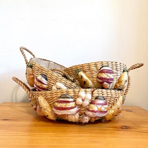 May include: Two woven wicker baskets stacked on a wooden surface. The baskets are decorated with straw eggs, garlic, and carrots. The baskets have rope handles and a burlap lining. The colors are natural, with red, white, and brown accents.