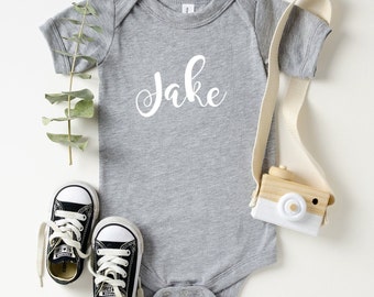 May include: Gray baby onesie with the name "Jake" written in white cursive lettering.