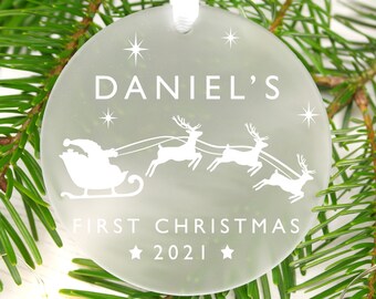 May include: White frosted glass Christmas ornament with a white silhouette of Santa Claus in a sleigh pulled by reindeer. The ornament says "Daniel's First Christmas 2021" and has small white stars.