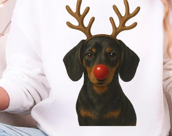 Op de afbeelding: Witte sweatshirt met een teckel met rendiergeweien en een rode neus. De afbeelding bevat een rood-witte mok met een peperkoekmannetje. Het thema is Kerstmis en vakantiekleding.