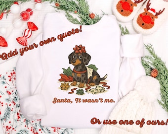 Peut inclure: Sweat-shirt blanc avec un dessin de teckel portant un nœud, entouré de papier cadeau et le texte "Santa, It wasn't me." L'image comprend un pyjama de Noël, un bonnet blanc et des pantoufles de renne.
