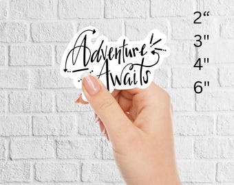 May include: A white sticker with the words "Adventure Awaits" in a stylish script font. The sticker is held against a white brick wall. Available sizes are 2", 3", 4", and 6 inches.