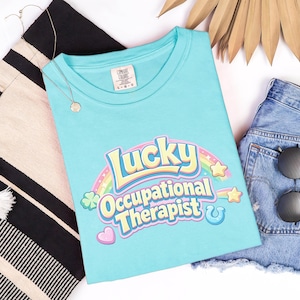 May include: A light blue t-shirt with the words "Lucky Occupational Therapist" in a colorful design. The shirt is paired with denim shorts, sunglasses, and a tote bag.