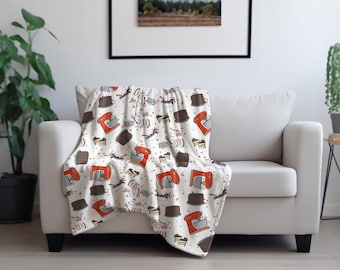 Puede incluir: Una manta blanca con un estampado de repostería, colocada sobre un sofá blanco. El estampado incluye batidoras rojas, pasteles de chocolate y otras ilustraciones de repostería. El sofá está contra una pared blanca con un cuadro enmarcado.