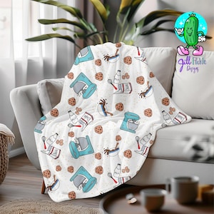 May include: A white throw blanket with a repeating pattern of baking-themed illustrations. The design includes light blue stand mixers, chocolate, milk cartons, eggs, and cookies. The blanket is draped over a gray sofa.