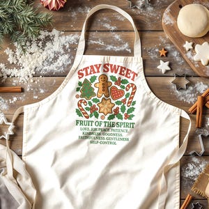 May include: Cream-coloured apron with the words "STAY SWEET" and "FRUIT OF THE SPIRIT" in red and green. The apron features gingerbread men, candy canes, and a Christmas tree design. Baking supplies and ingredients are scattered around the apron.