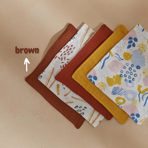 Könnte beinhalten: Eine Sammlung von fünf quadratischen Stoffservietten in verschiedenen Farben und Mustern. Die Servietten umfassen einfarbiges Braun, Rost und Gelb sowie zwei gemusterte Servietten mit abstrakten Designs in Braun, Blau und Rosa auf weißem Hintergrund.