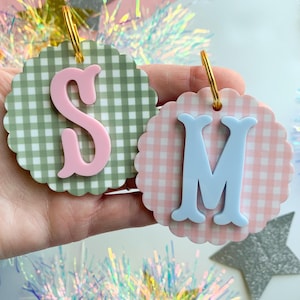 May include: Two decorative round ornaments with gold hoops. One has a pink letter "S" on a green and white gingham background. The other has a light blue letter "M" on a pink and white gingham background. Both have scalloped edges.