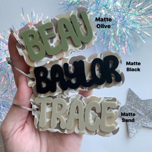 May include: Three personalized name tags with a camouflage pattern background. The names "BEAU" in matte olive, "BAYLOR" in matte black, and "TRACE" in matte sand are layered on top. Each tag has a silver-colored metal ring.