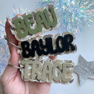 May include: Three personalized name tags with a camouflage pattern background. The names "BEAU" in olive green, "BAYLOR" in black, and "TRACE" in beige are layered on top. Each tag has a silver keyring.