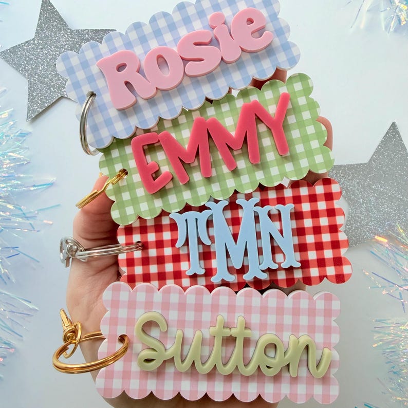 May include: Four rectangular acrylic keychains with scalloped edges and gingham patterns in blue, green, red, and pink. Each keychain features raised 3D lettering in contrasting colors displaying the names Rosie, Emmy, TMN, and Sutton.