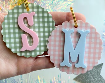 May include: Two circular scalloped tags with gold metal rings. One tag features a sage green gingham pattern with a raised pink letter S. The other tag features a light pink gingham pattern with a raised light blue letter M on the front.