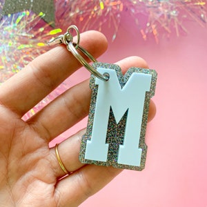 May include: A white letter "M" keychain with a silver glitter border and a silver keyring. The keychain is held against a pink background. The letter has a collegiate style.