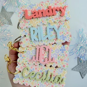 May include: Four rectangular acrylic tags with scalloped edges and a colorful floral pattern. Each tag features a raised name or monogram: Landry in coral, RILEY in light blue, a pink monogram, and Cecilia in light green. Each tag has a metal ring.