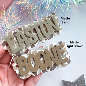 May include: Two custom name hair clips. The top clip is matte sand with the name "EASTON" in a 3D design. The bottom clip is matte light brown with the name "BOONE". Both clips have a camouflage pattern.