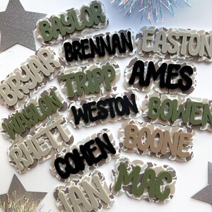 May include: A collection of personalized name tags with a camouflage pattern background. The names are in various colors, including green, black, and tan. The names are in a bold, rounded font.